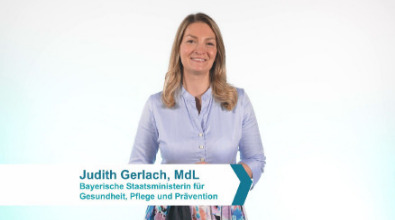 Eine blonde Frau steht vor einer hellen Wand und spricht in die Kamera.